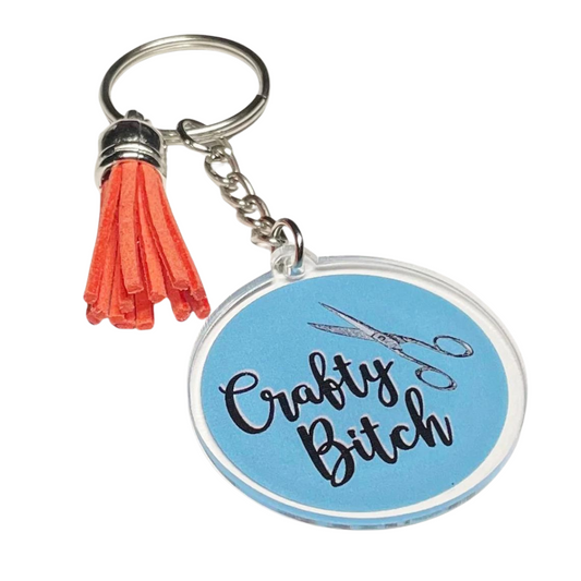 A photo a cute keychain. It's round and blue. On it, there's pair of scissors. Text on keychain reads 'Crafty Bitch.'