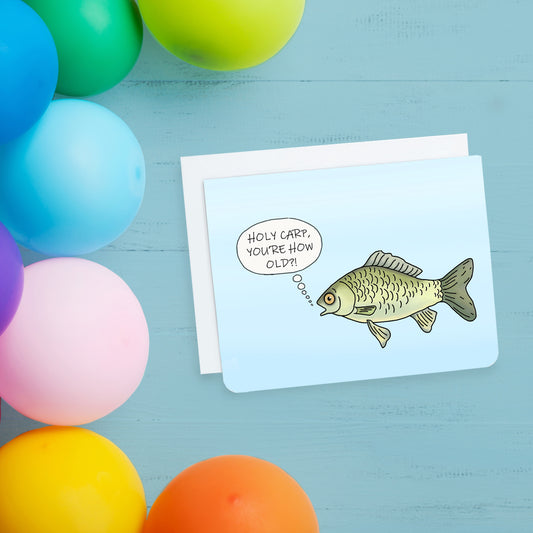 A photo of a blue birthday card. It has a surprised looking cartoon fish on it. It has a speech bubble which says, 'Holy carp. You're how old?!'