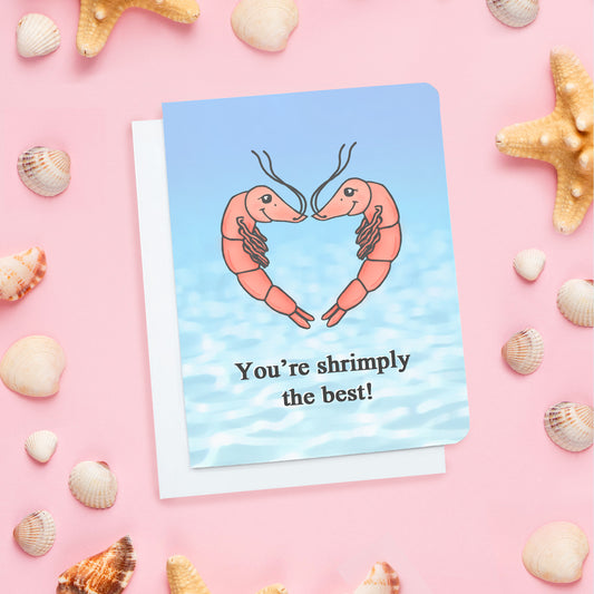 A cute card. It is blue and has two shrimp on it smiling at one another in a heart shaped formation. Text blow on card reads: 'You're shrimply the best.'