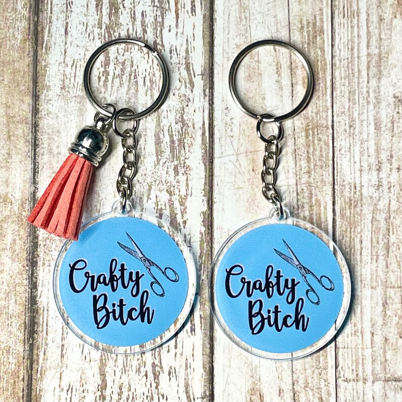 A photo of two cute keychains. They're round and blue. On them, there's pair of scissors. Text on keychain reads 'Crafty Bitch.'