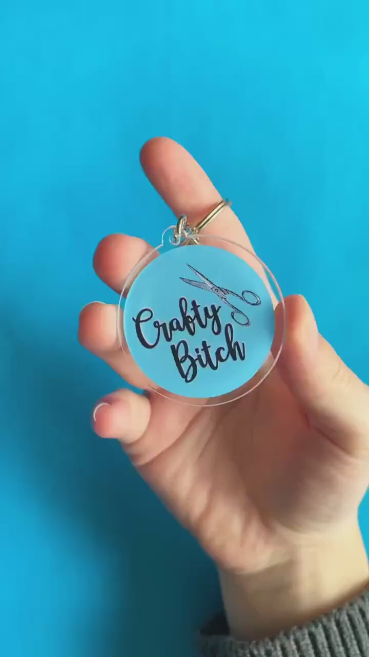 A video of someone showing off a key chain. It's round and blue. On it, there's pair of scissors. Text on keychain reads 'Crafty Bitch.'