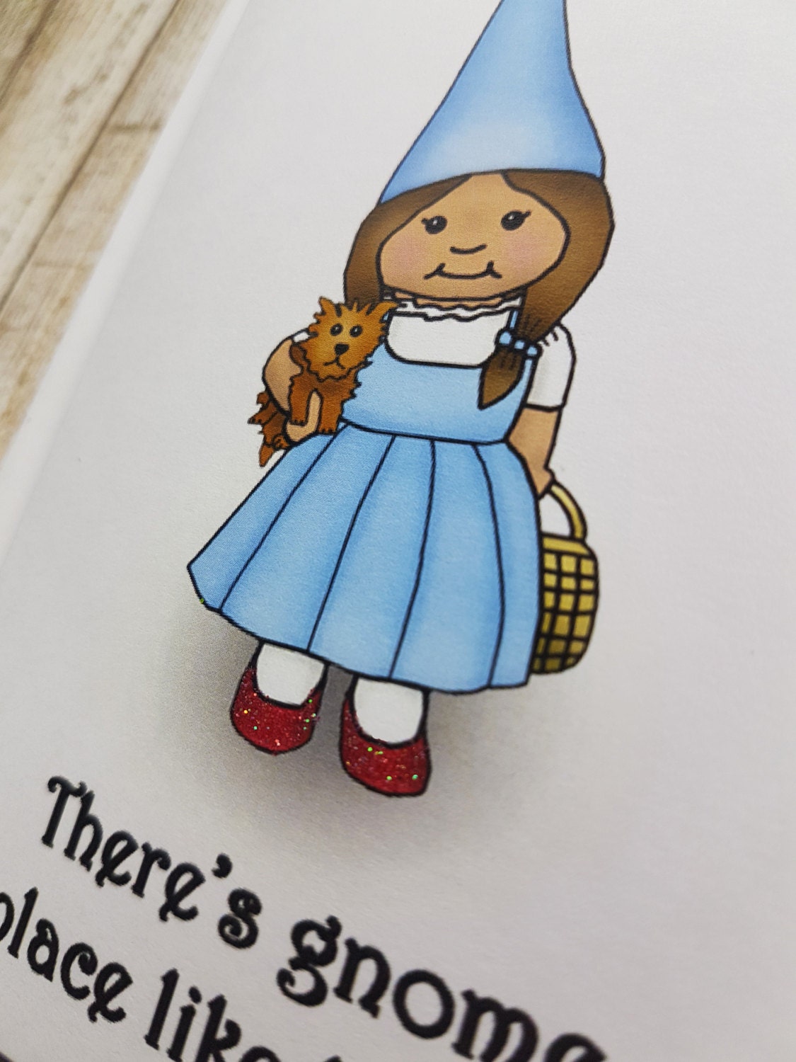 A photo of a greeting card. It has a cartoon gnome on it dressed like Dorothy from the Wizard of Oz holding a little dog. Text reads, There's Gnome Place Like Gnome.