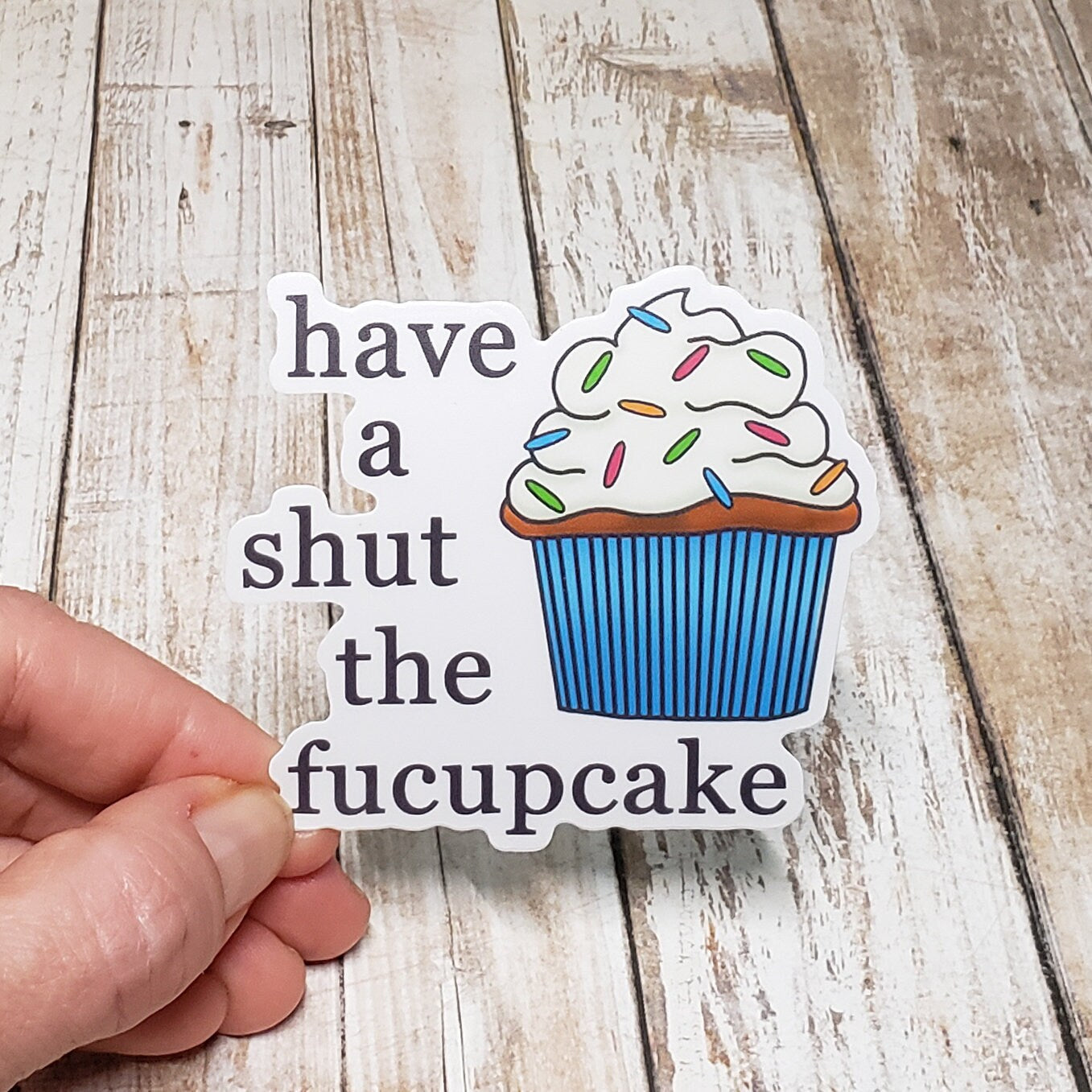 A photo of a vinyl sticker of cartoon cupcake. Text on funny sticker reads 'have a shut the fucupcake.'