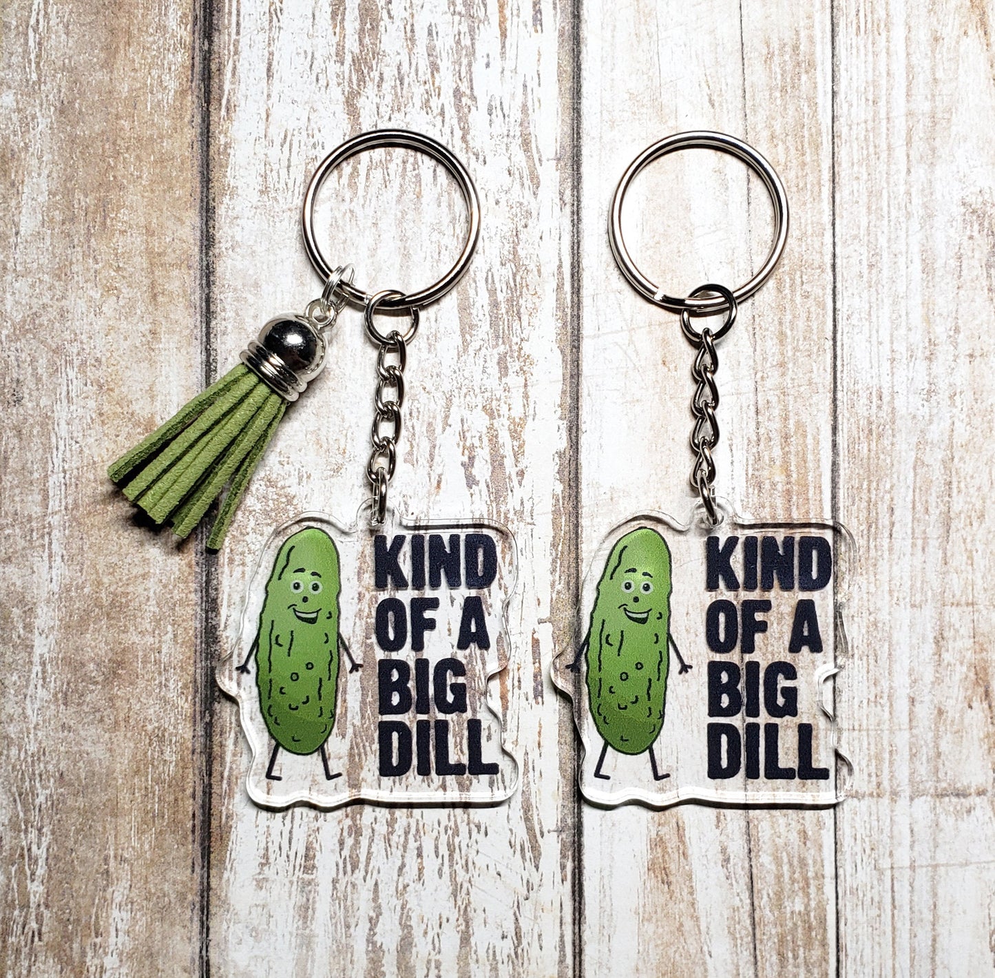 A photo of two acrylic keychains. They have smiling pickles on them. Text on keychain reads 'Kind of a Big Dill.'