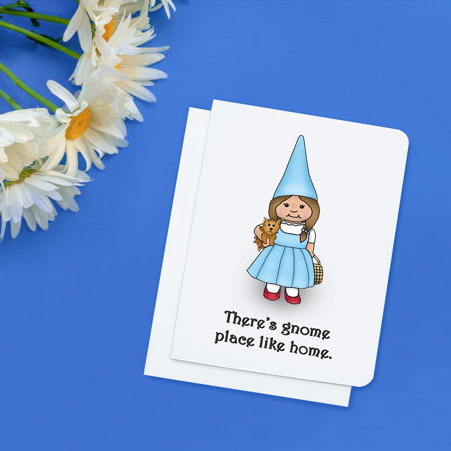 A photo of a greeting card. It has a cartoon gnome on it dressed like Dorothy from the Wizard of Oz holding a little dog. Text reads, There's Gnome Place Like Gnome.