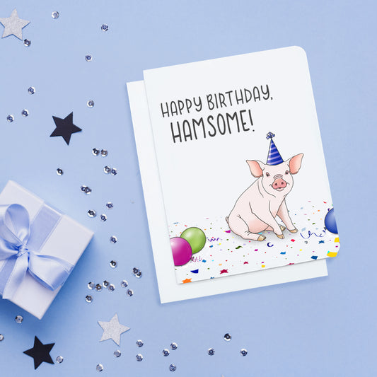 A photo of a white birthday card. It has a cartoon pig sitting down wearing a party hat. There are balloons on the floor. Text on card reads, 'Happy Birthday, Hamsome!'