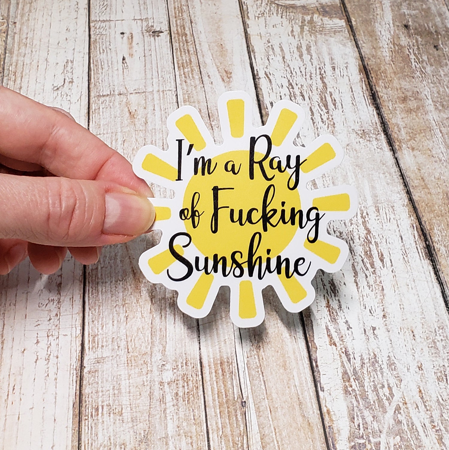 Vinyl Sticker with graphic yellow sunshine on white background with text reading "I'm a Ray of Fucking Sunshine" in black cursive font.