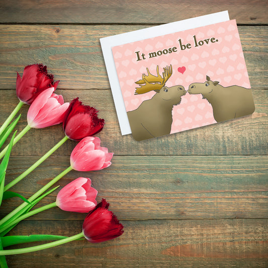 A photo of a pink valentines card. It has two cartoon moose on it kissing. Text on card reads 'It moose be love.'