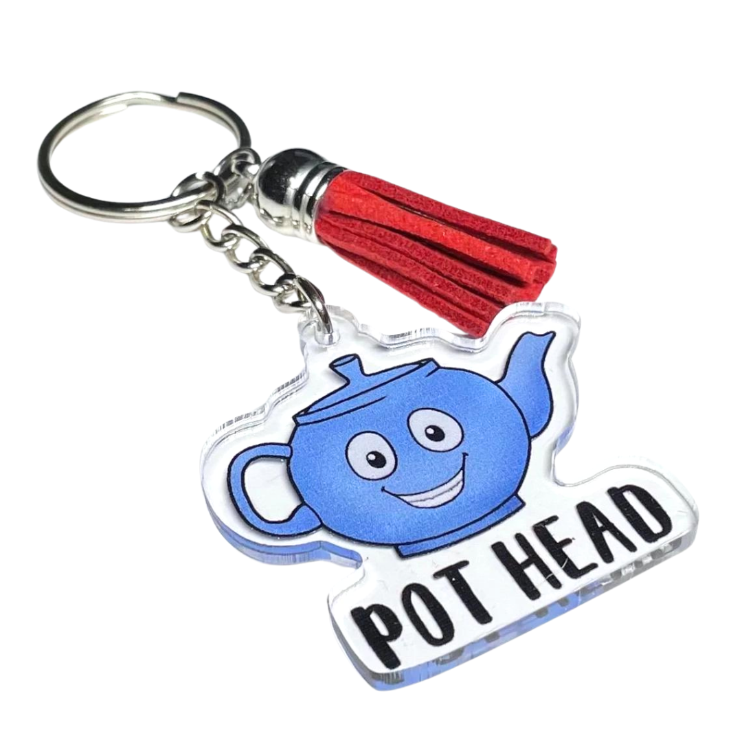 A photo of a cute keychain. It has a blue cartoon teapot on it; it's smiling. Text on keychain reads, Pot Head.