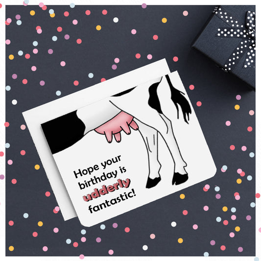 A photo of a white birthday card. It has the underside of a cartoon cow on it. Text on card reads, 'Hope Your Birthday is Udderly Fantastic!'