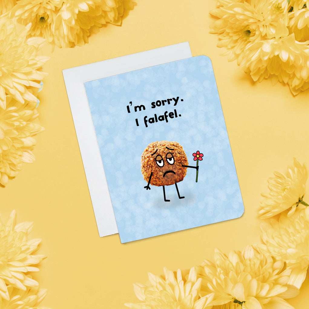 A photo of a blue apology card. It has a photo of a falafel on it. It has cartoon stick arms and legs on it and sad cartoon eyes. It's holding a red flower. Text on card reads, 'I'm sorry. I falafel.'