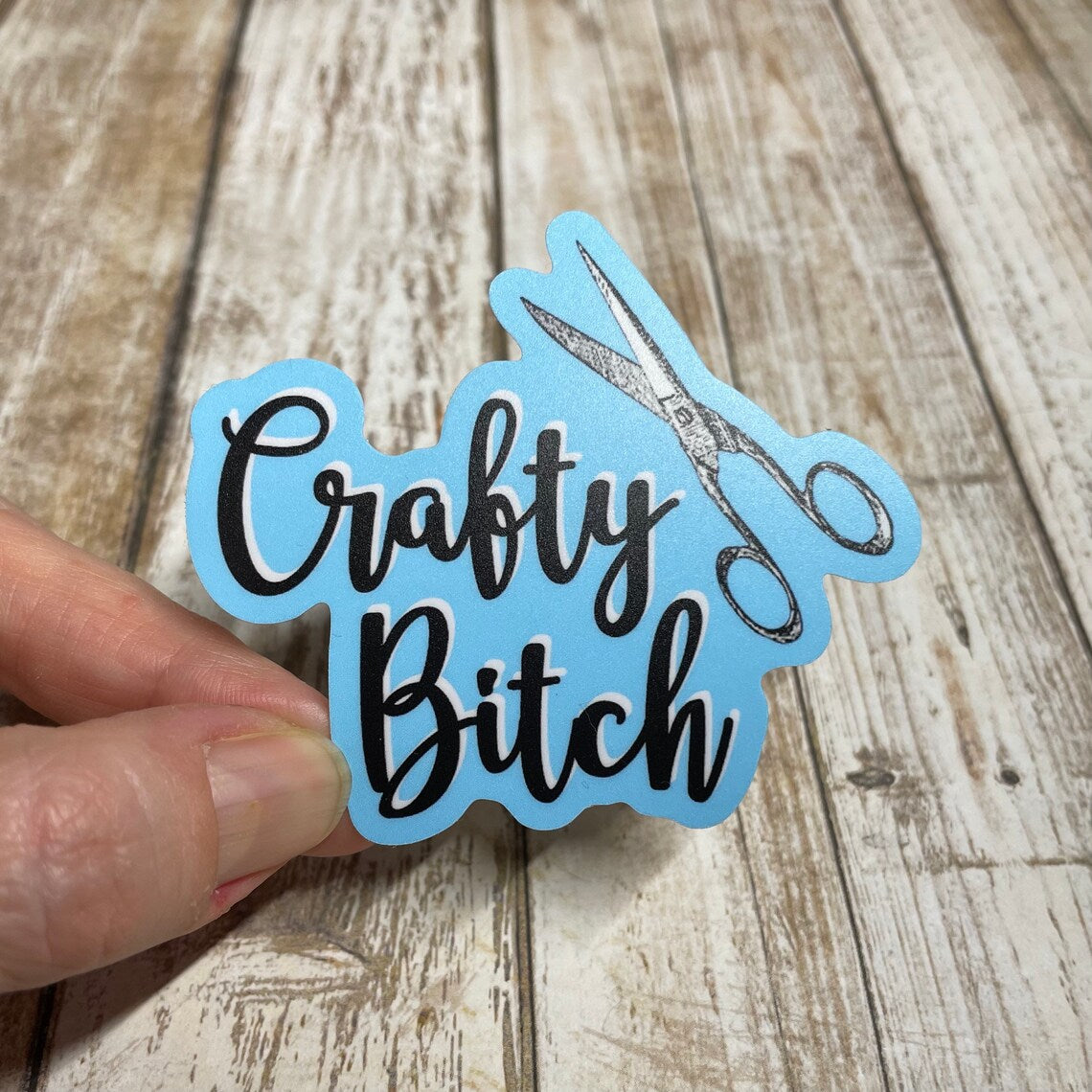 A photo of a blue cute sticker. It says 'Crafty Bitch' on it and there's a pair of scissors on the sticker.