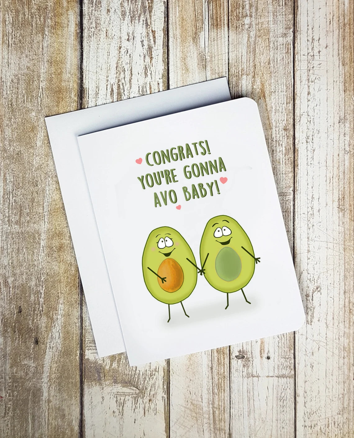 A photo of a white baby shower card. It has two halves cartoon smiling avocados on it. One side has the stone.  Text on card reads, 'Congrats! You're Gonna Avo Baby!'