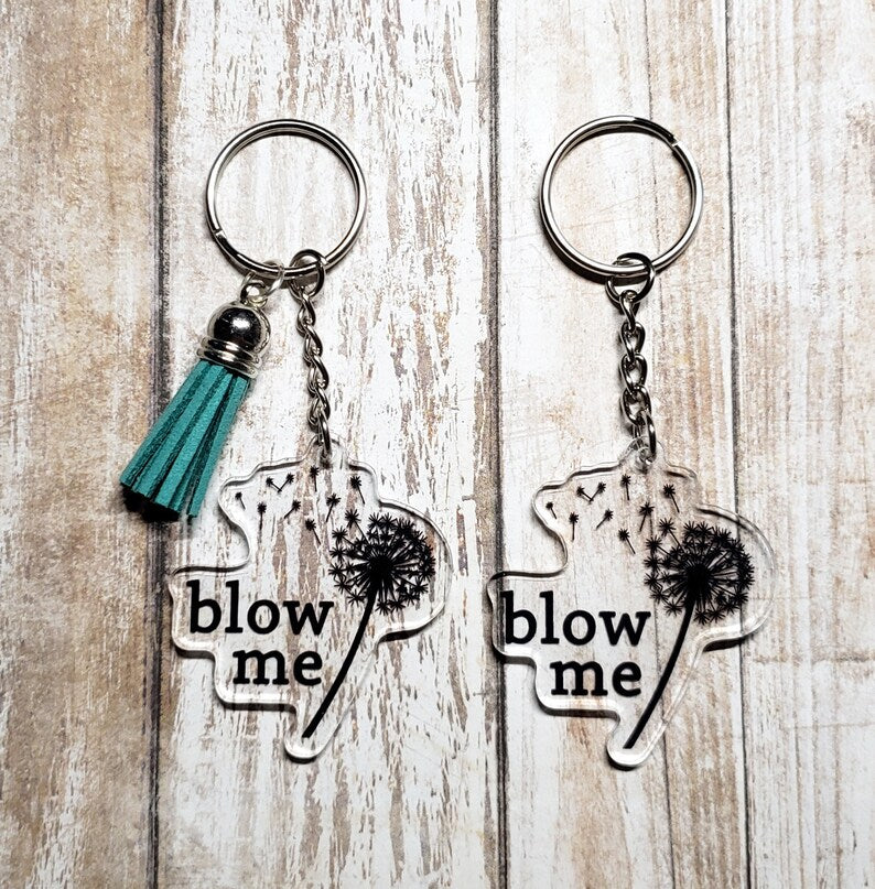 A photo of two key chains. They have blown dandelions on them. Text on key chain reads 'Blow Me.'