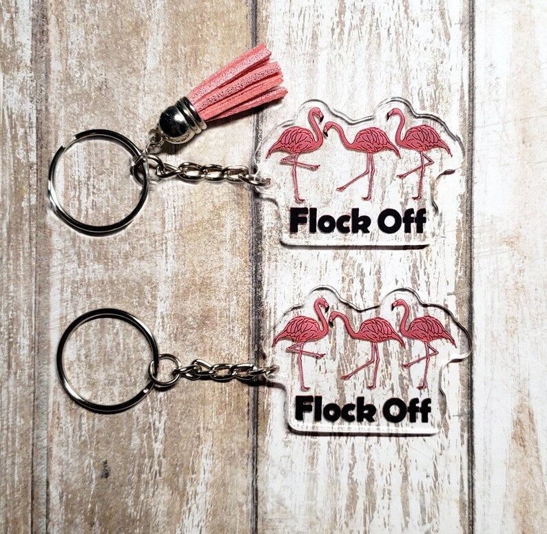 A photo of two key chains. They have three flamingoes on them. Text below flamingos reads "Flock Off.'