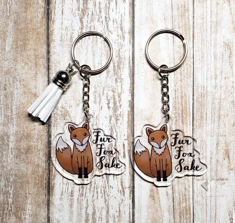 A photo of two key chains. The keychains have cartoon foxes on them. Text on keychain reads 'Fur Fox Sake.'