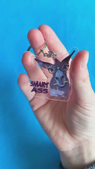 A video of a hand showing off an acrylic keychain with a donkey on it. The donkey has a pencil in its mouth and glasses on. Text on keychain reads 'Smart Ass.'