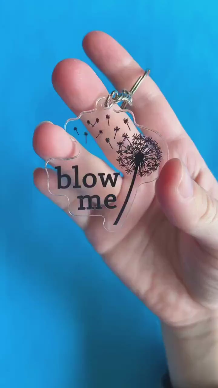 A video of a hand showing off a key chain. It has a blown dandelion on it. Text on key chain reads 'Blow Me.'