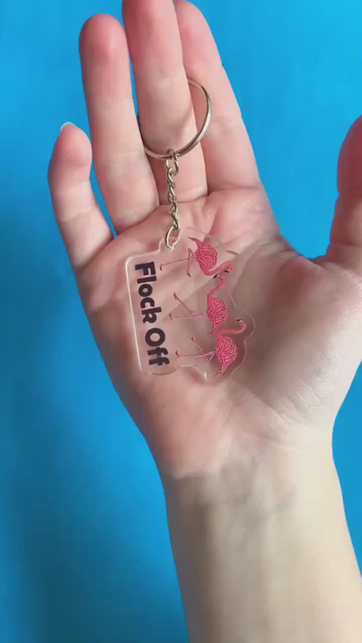 A video of a hand displaying a key chain. It has three flamingoes on it. Text below flamingos reads "Flock Off.'