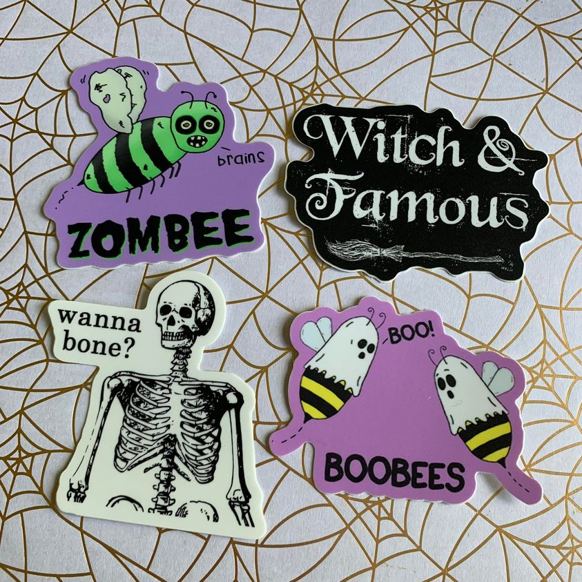 A photo of 4 halloween stickers. 1 a zombie bee text on sticker reads 'Zombee.' 2 a broom stick text reads 'Witch & Famous.' 3 a skeleton text reads 'Wanna Bone?' and 4 two bees with sheets on their heads text reads 'BooBees.'