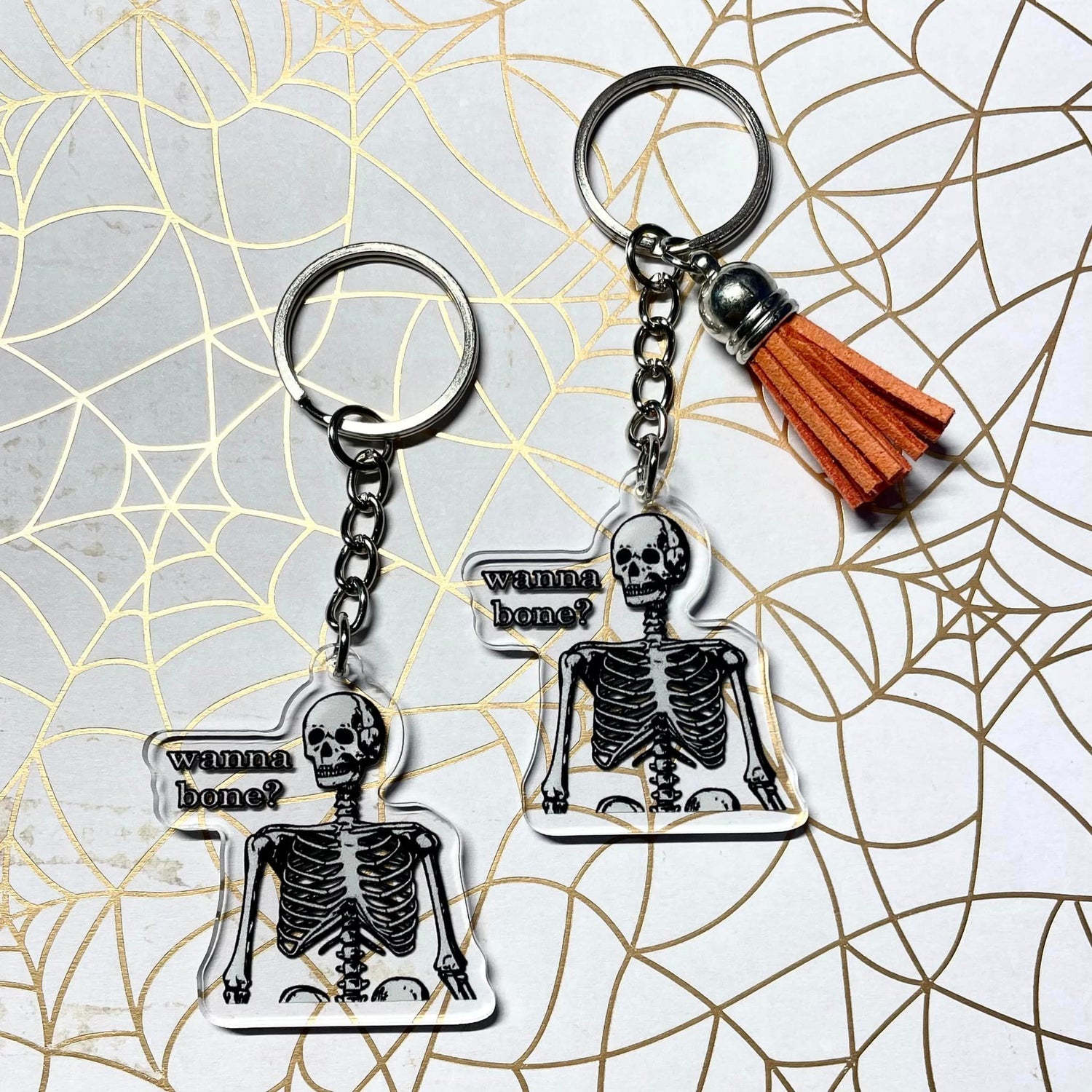 A photo of two halloween keychains. The keychain has a skeleton on it. Text on keychain reads 'Wanna Bone?'