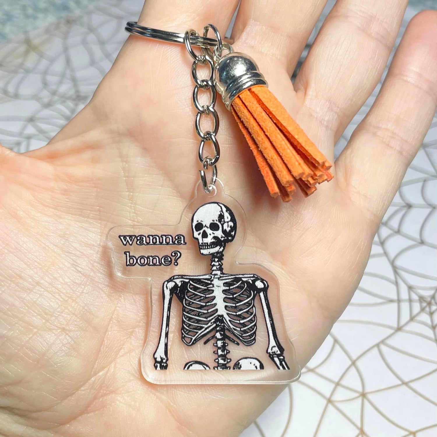 A photo of a halloween keychain. The keychain has a skeleton on it. Text on keychain reads 'Wanna Bone?'
