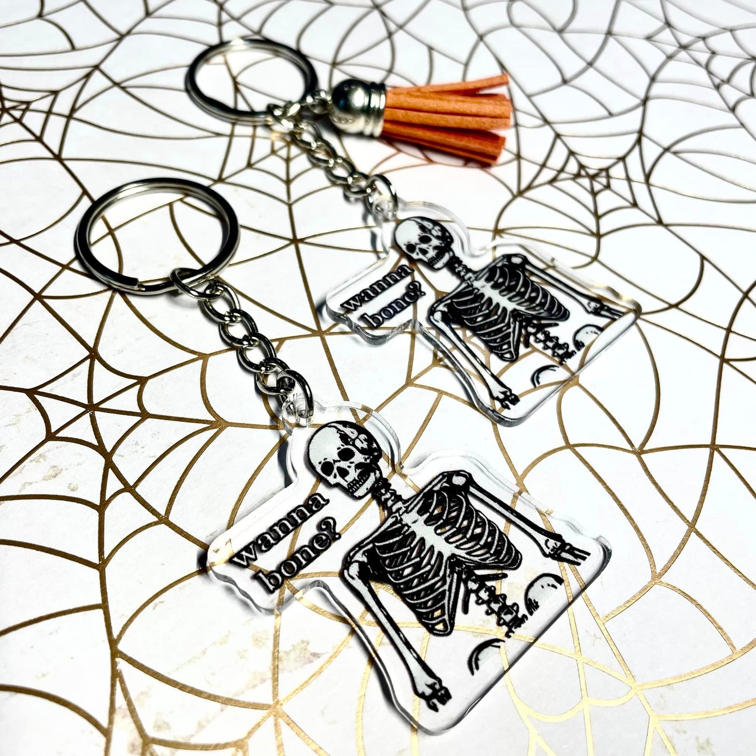 A photo of two halloween keychains. The keychain has a skeleton on it. Text on keychain reads 'Wanna Bone?'