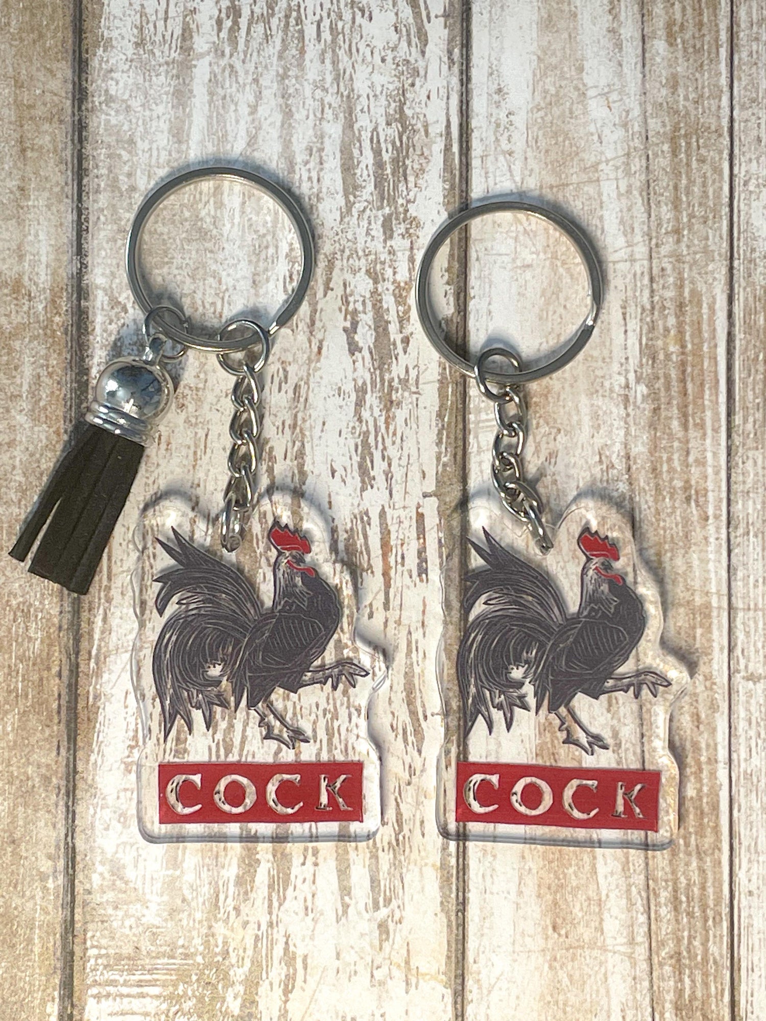 A photo of two acrylic key chains. They have black cockerels on them. Text below bird reads 'COCK.'