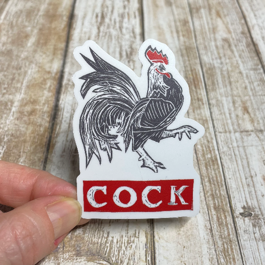 A photo of a sticker. It has a strutting cockerel on it. Text on sticker reads 'Cock.'
