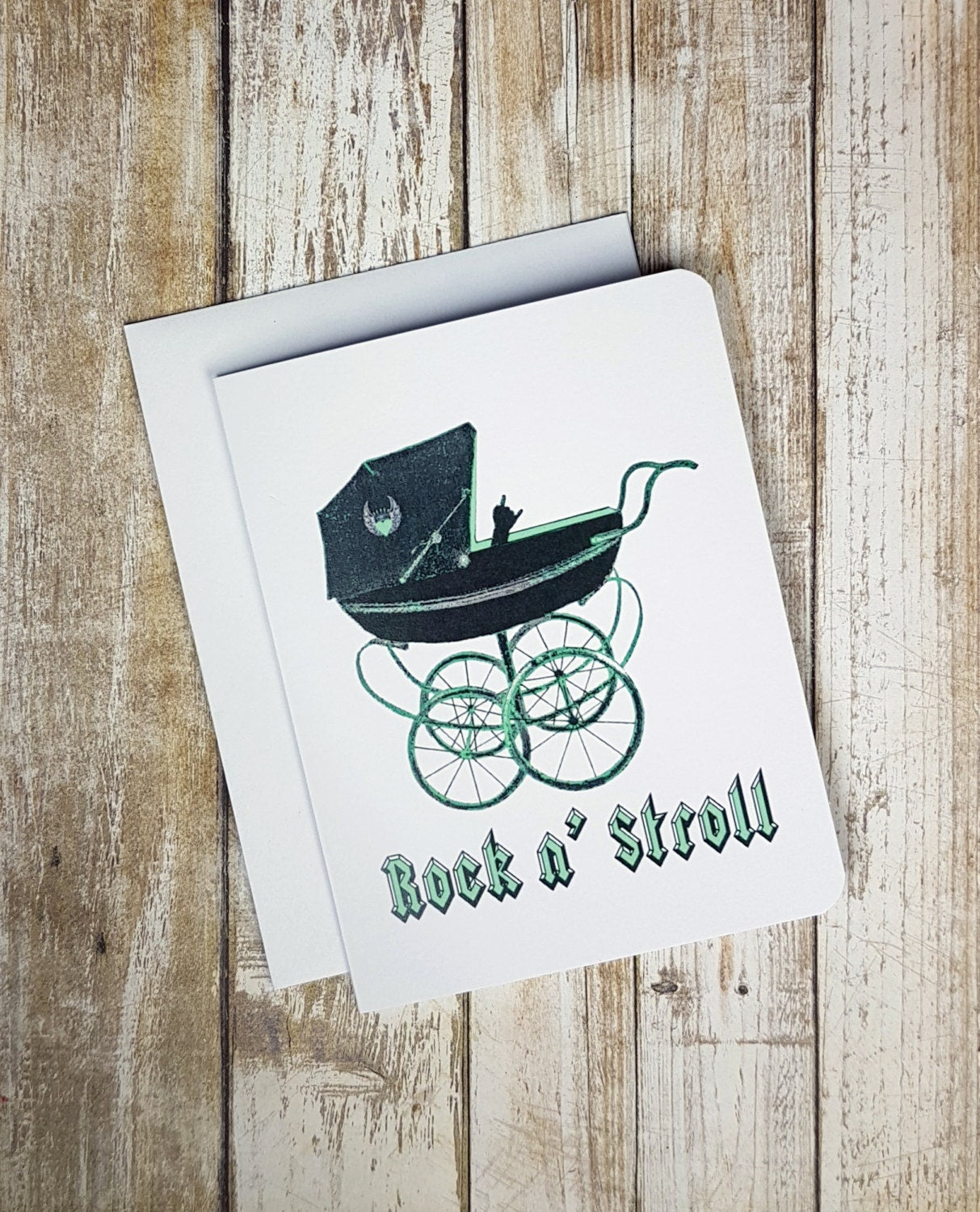 A photo of a white baby shower card. It has a graphic, vintage baby stroller on it. There's a little baby hand peeking out making the rock symbol. Text on card reads, 'Rock N' Stroll.'