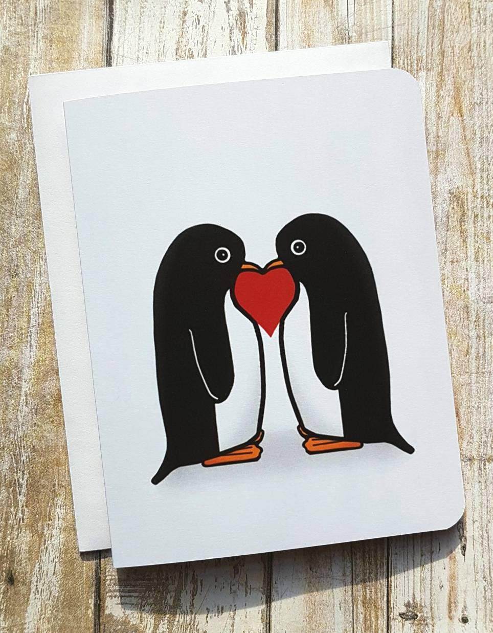 A photo of a cute card. It has two cartoon penguins on it facing each other. There's a red love heart between them.