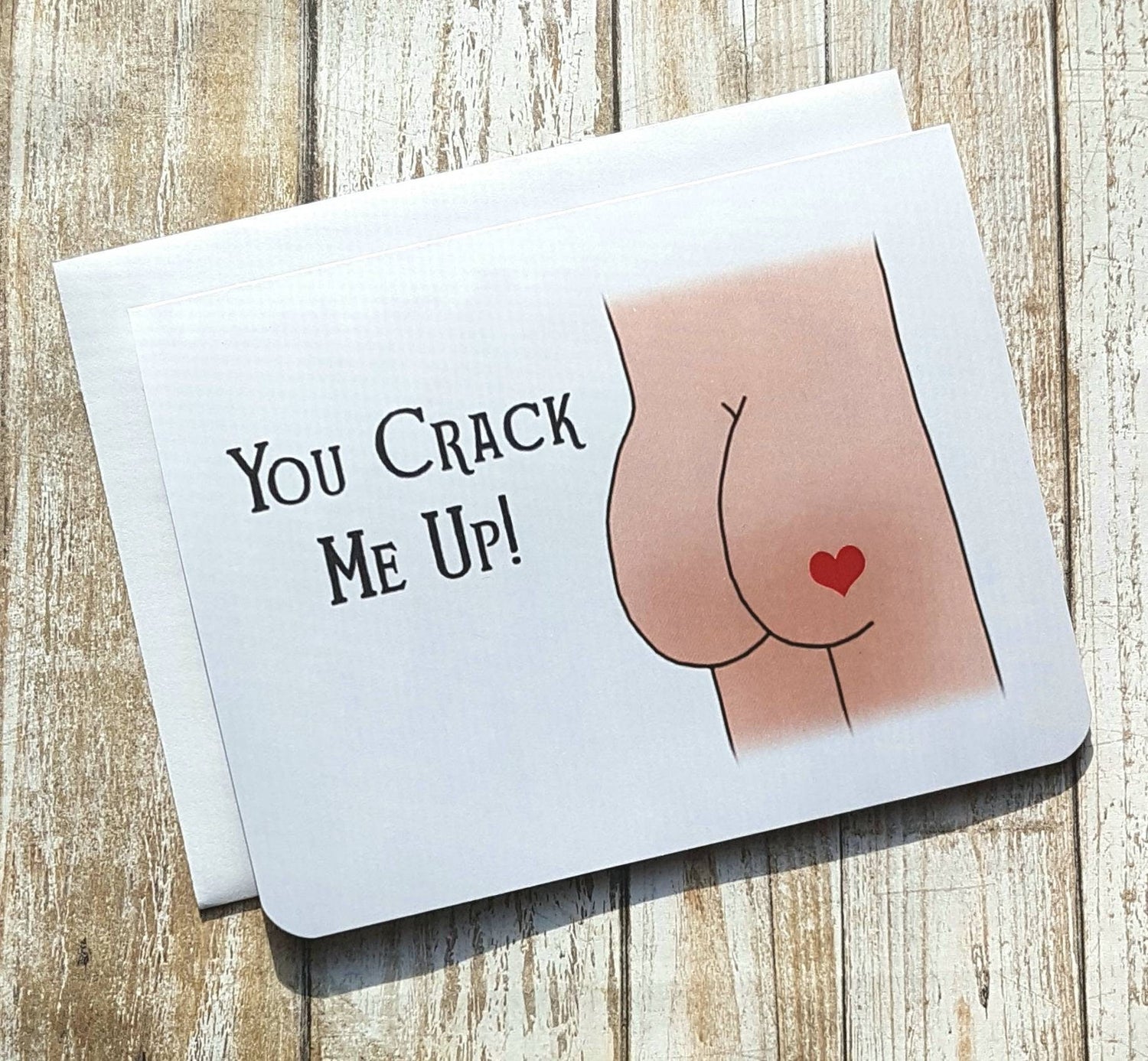 A photo of a white greeting card. It has a cartoon butt on it. The one butt cheek has a red love heart on it. Text on card reads 'You Crack Me Up.'