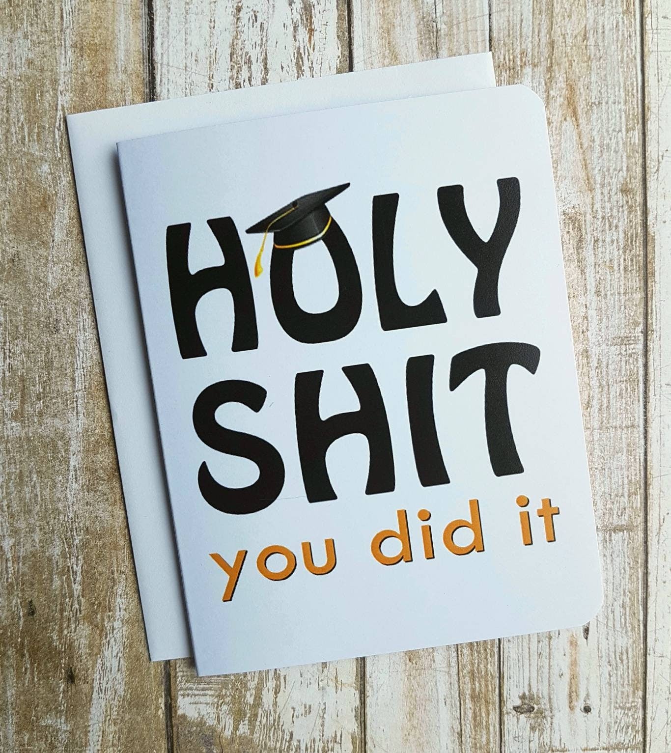 A photo of a white graduation card. In bold text it reads, 'Holy Shit. You did it!' the 'O' on holy is wearing a graduation cap.