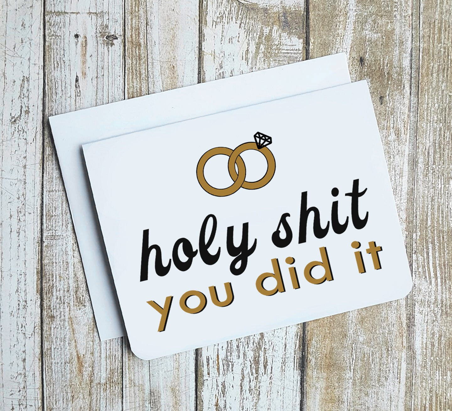 A photo of a white wedding card. It has two rings intersecting on it. Text on card reads, 'Holy Shit you did it.'