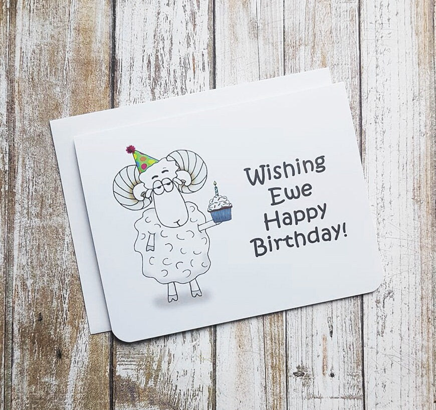 A photo of a white birthday card. It has a cartoon sheep on it. It's holding a cupcake. Text on card reads, 'Wishing Ewe Happy Birthday.'