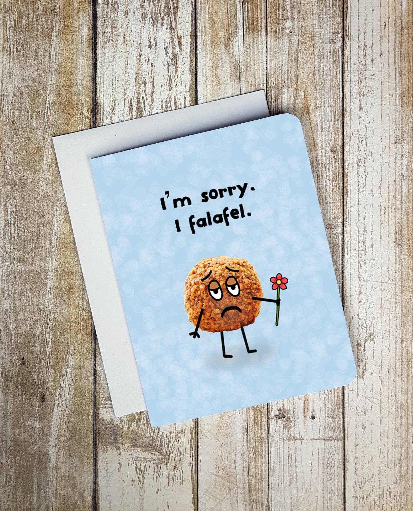 A photo of a blue apology card. It has a photo of a falafel on it. It has cartoon stick arms and legs on it and sad cartoon eyes. It's holding a red flower. Text on card reads, 'I'm sorry. I falafel.'