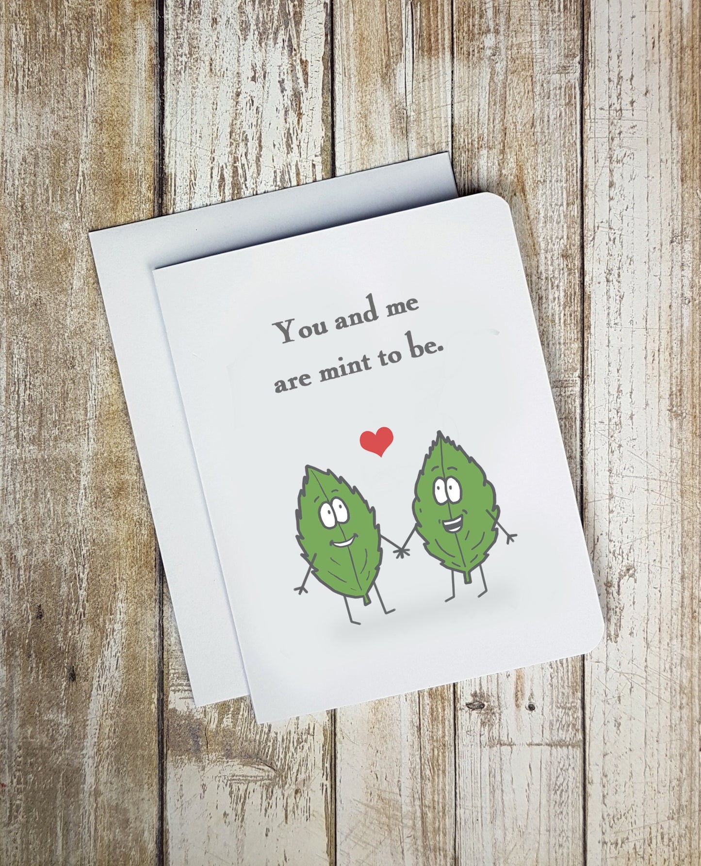 A photo of a love card. It has two cartoon mint leaves on it. They're smiling at one another. There's a love heart between them. Text reads 'You and Me Are Mint to Be.'