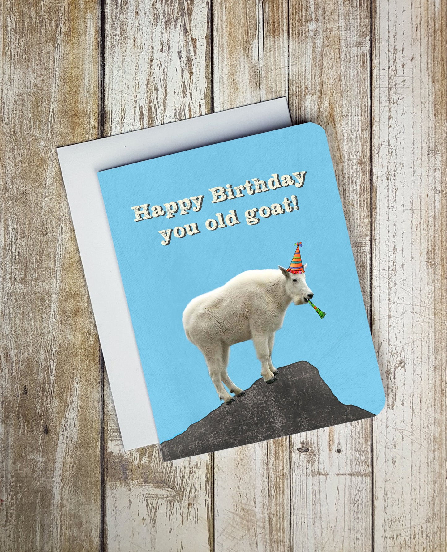 A photo of a blue birthday card. It has a photo of a mountain goat on it. It's wearing a party hat and has a streamer in its mouth. Text on card reads, 'Happy Birthday you old goat!'