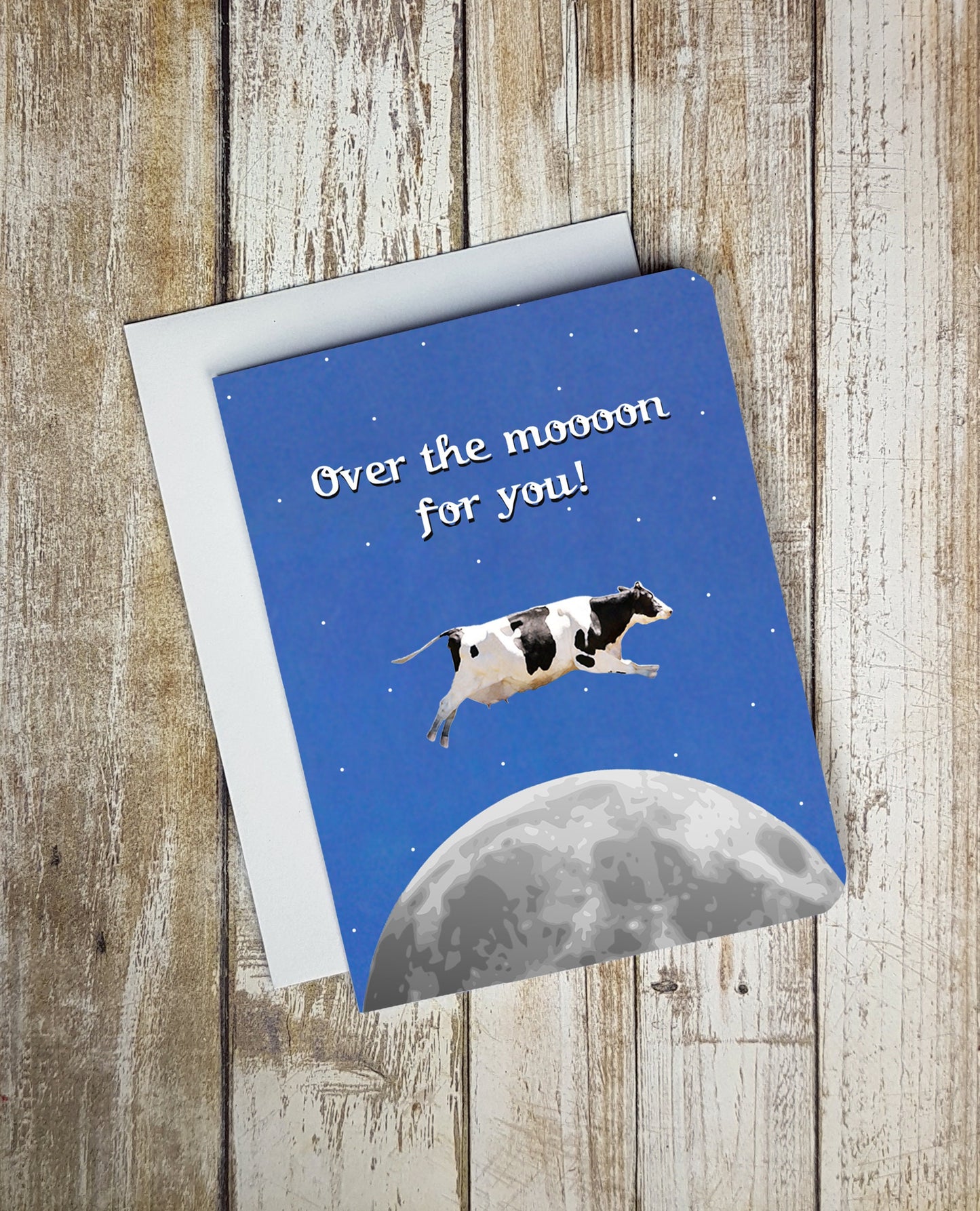 A photo of a blue greeting card. It has a cow jumping one the moon. Text on card reads, 'Over the moooon for you!'
