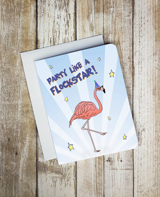 A photo of a white and blue birthday card. It has a cartoon flamingo on it. Its wearing a party hat and sunglasses. Text on card reads, 'Party Like A Flockstar.'