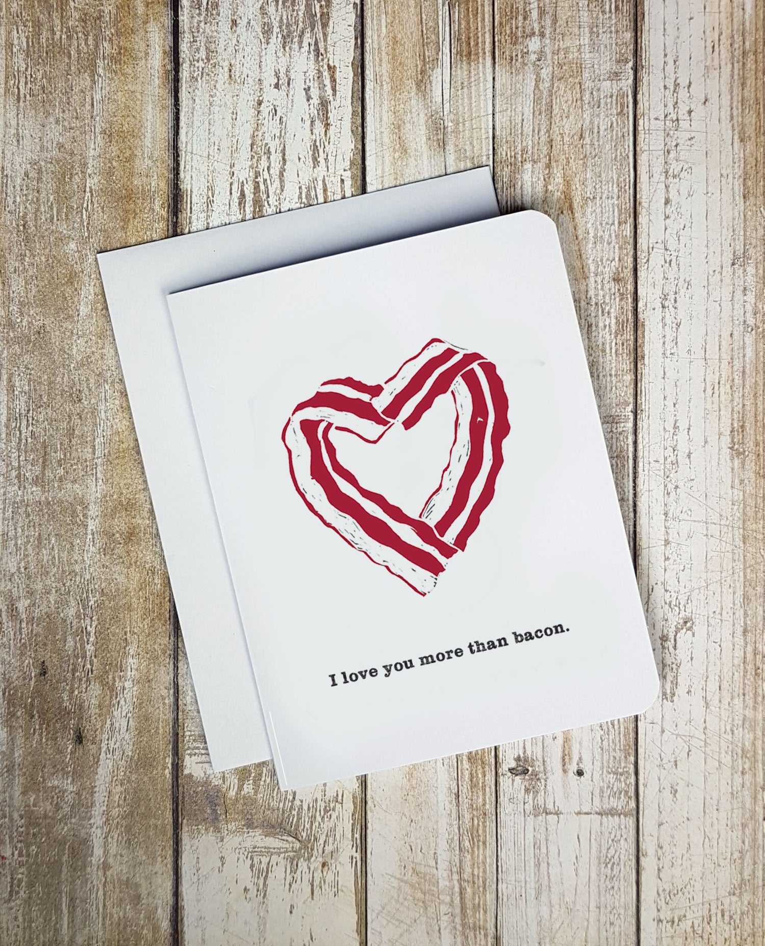 A photo of a white valentine's card. It has cartoon bacon on it in a love heart formation. Text on card reads, 'I love you more than bacon.'