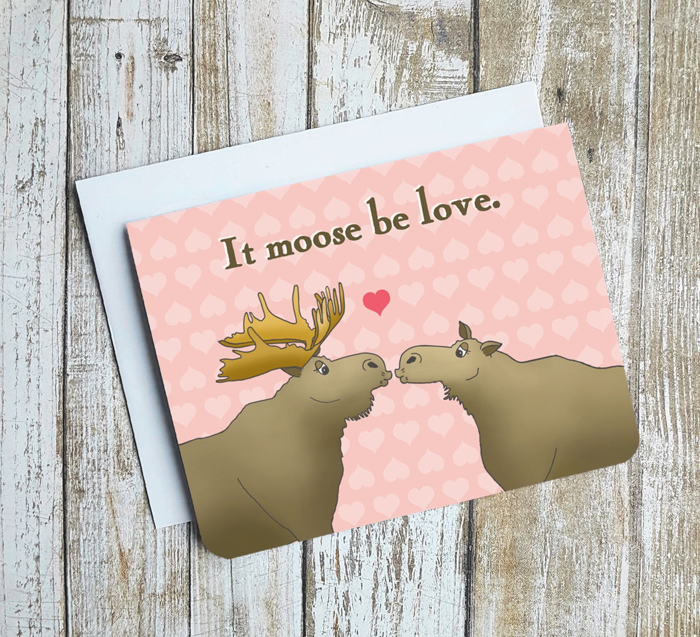 A photo of a pink valentines card. It has two cartoon moose on it kissing. Text on card reads 'It moose be love.'