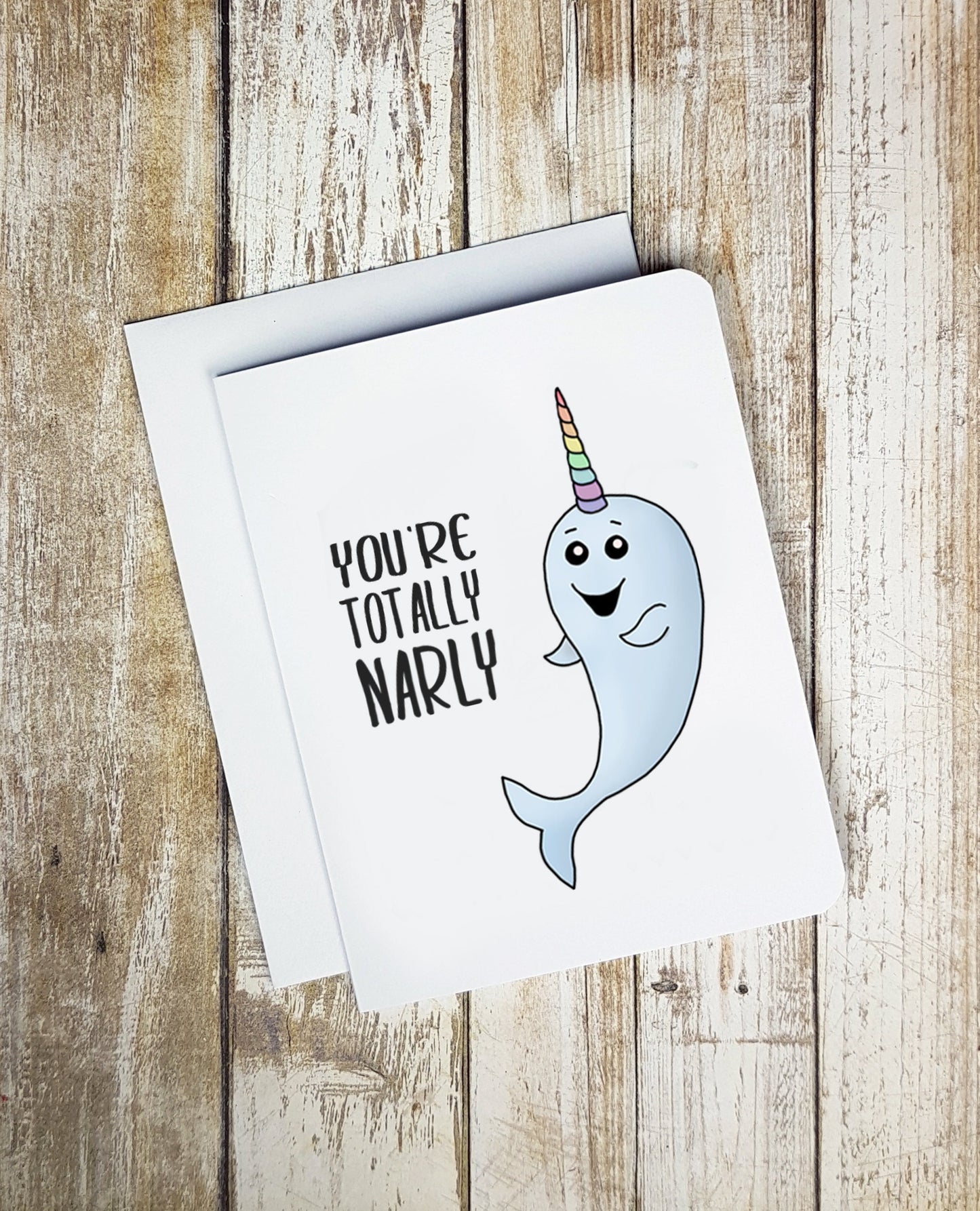 A cute white greeting card. It has a smiling cartoon narwhal on it with a striped colourful horn. Text on card reads 'You're Totally Narly.'