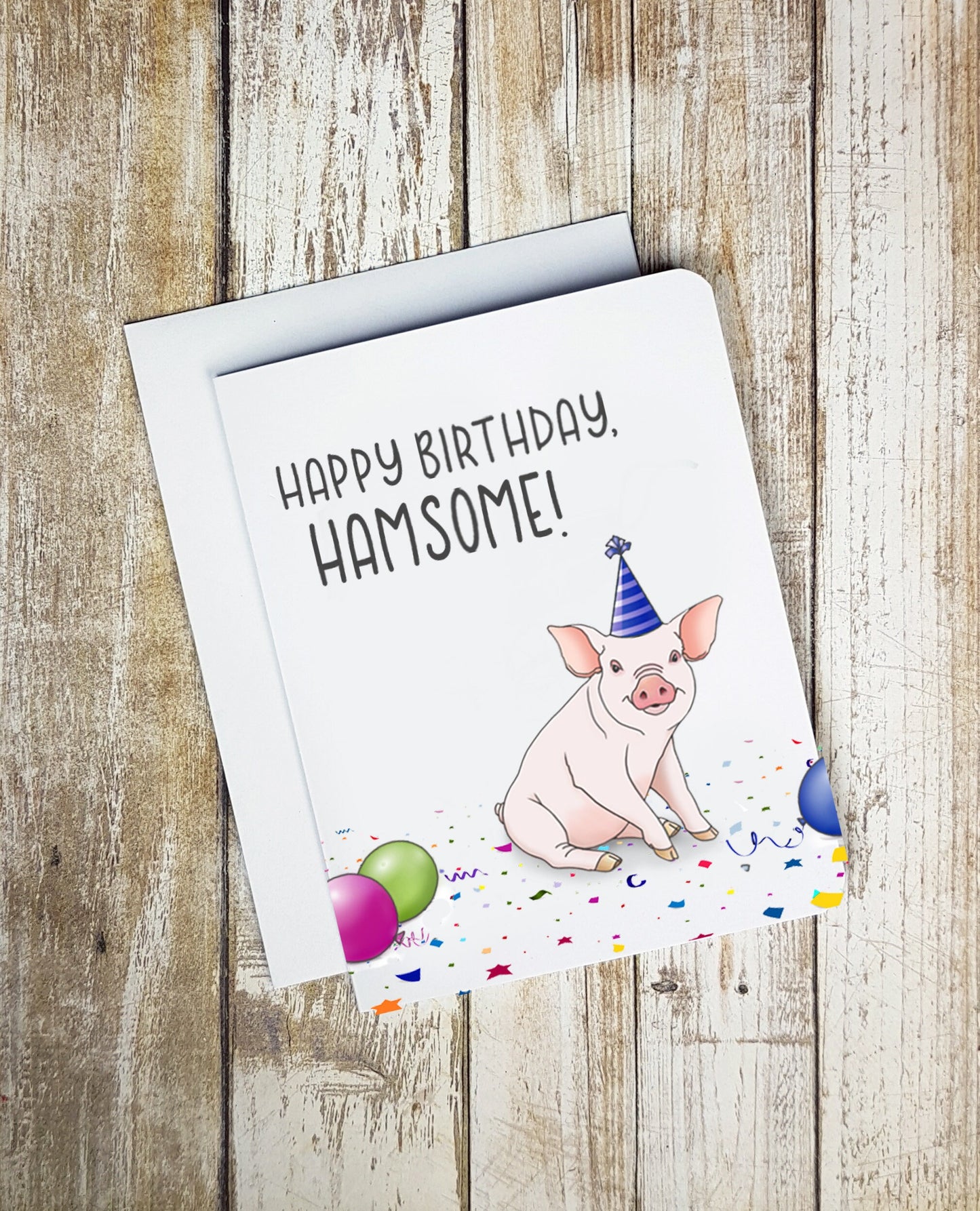 A photo of a white birthday card. It has a cartoon pig sitting down wearing a party hat. There are balloons on the floor. Text on card reads, 'Happy Birthday, Hamsome!'