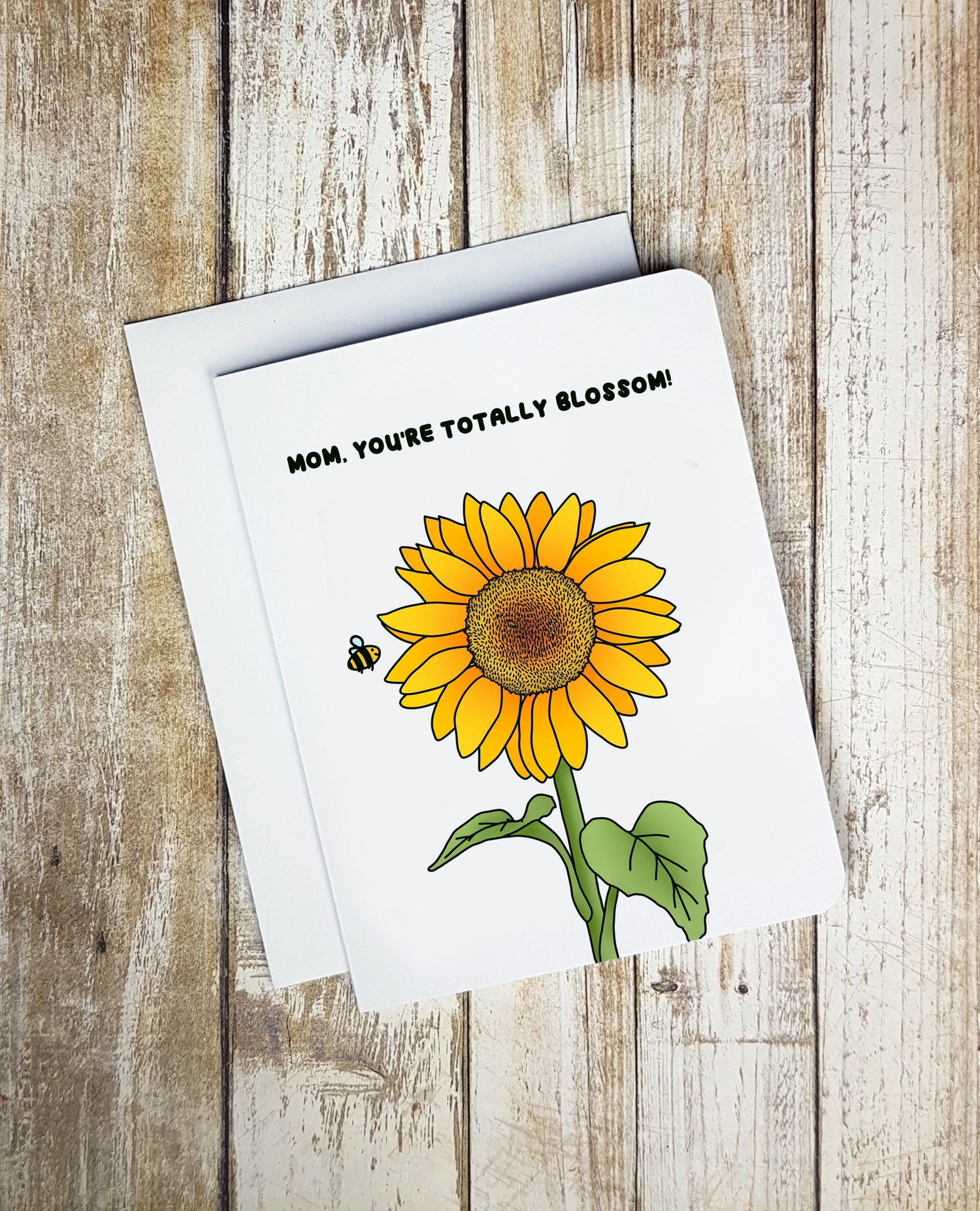 A photo of a mothers day card. It has a sunflower and a bee on it. Text reads, 'Mom, You're Totally Blossom!'