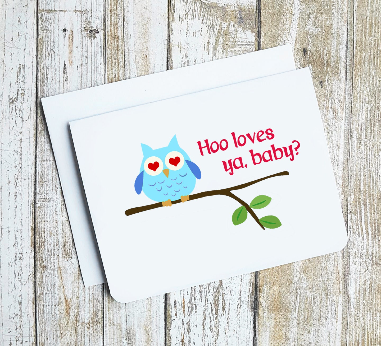 A photo of a white valentines card. It has an cartoon owl on a branch. It has love hearts as pupils. Text on card reads, 'Hoo loves ya, baby?'