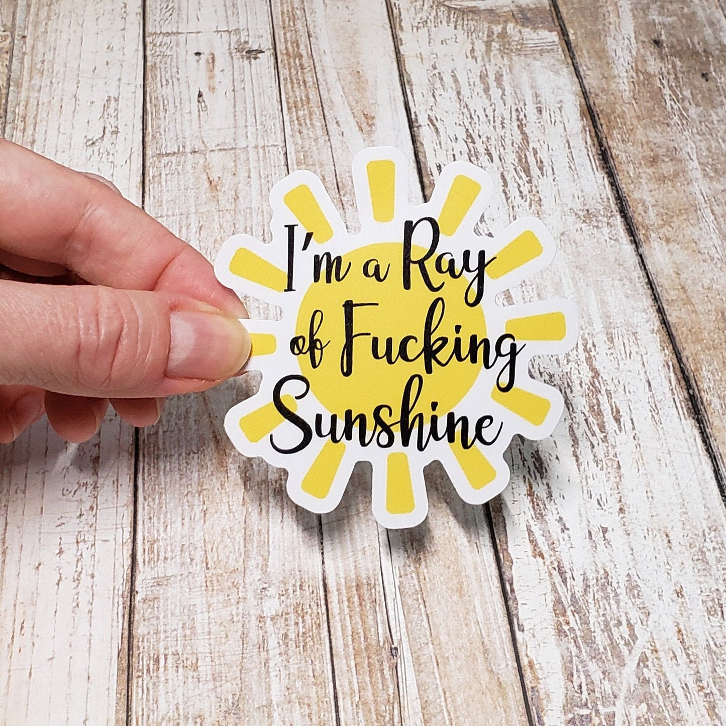 A photo of a vinyl sticker of a yellow sunshine. Text on sticker reads 'I'm a ray of fucking sunshine.'