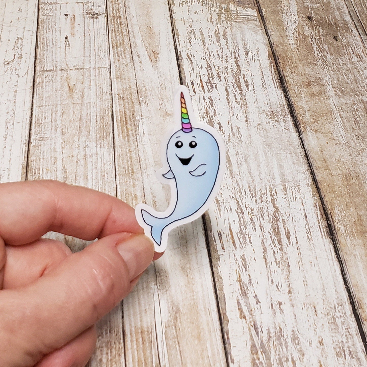 A photo of a cute sticker. It has a smiling narwhal on it. Its horn is colourful, like a unicorn.