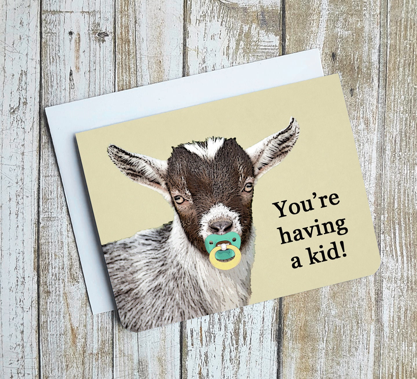 A photo of a baby shower card. It has a cartoon goat on it. It has a pacifier in its mouth. Text on card reads, 'You're having a kid!'
