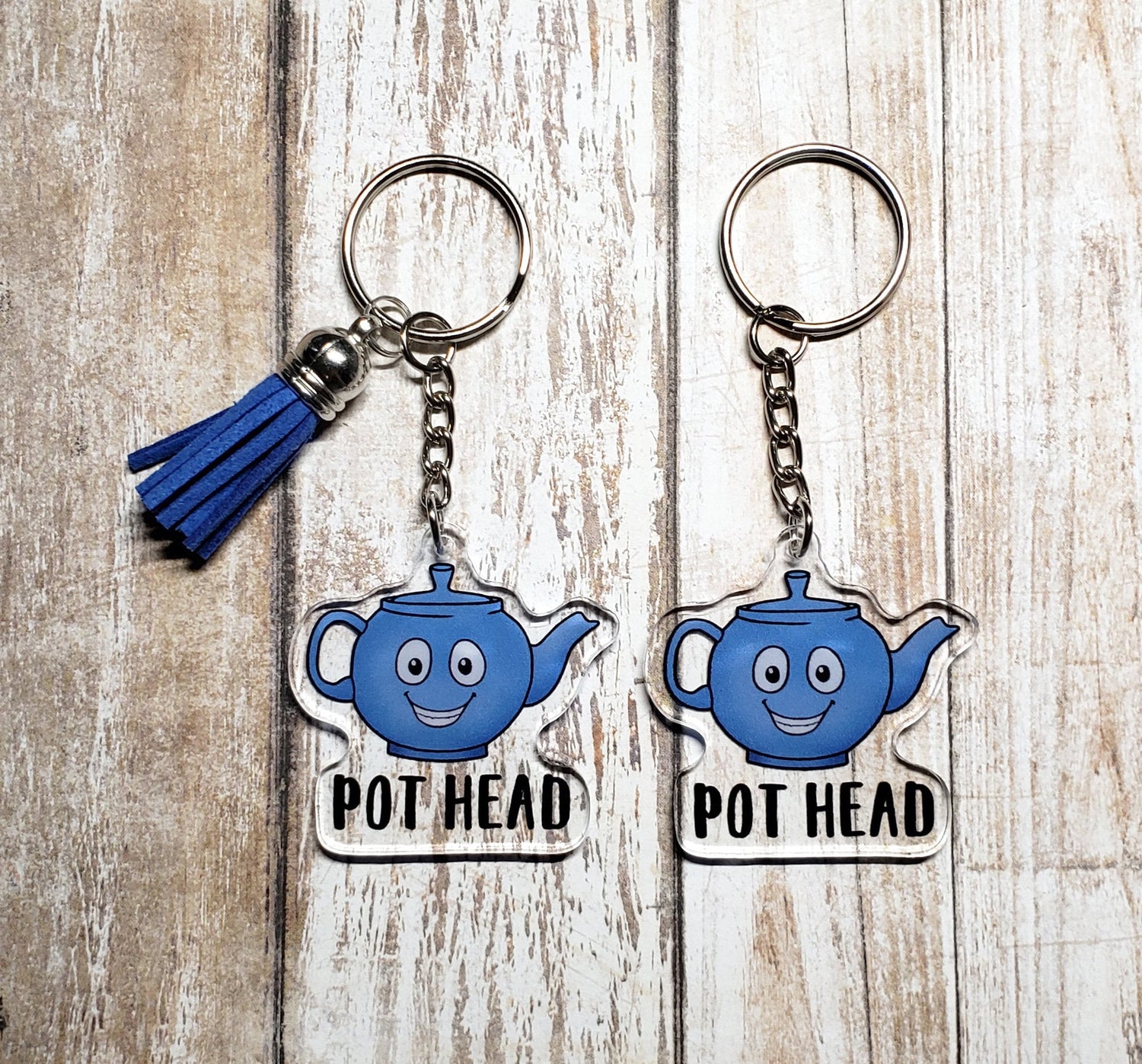 A photo of two acrylic key chains. The keychains have smiling blue teapots on them. Text on keychain reads 'Pot Head.'